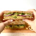 View of the cut side of a avocado chick pea sandwich. There are hands on each side of the sandwich presenting it to the camera.