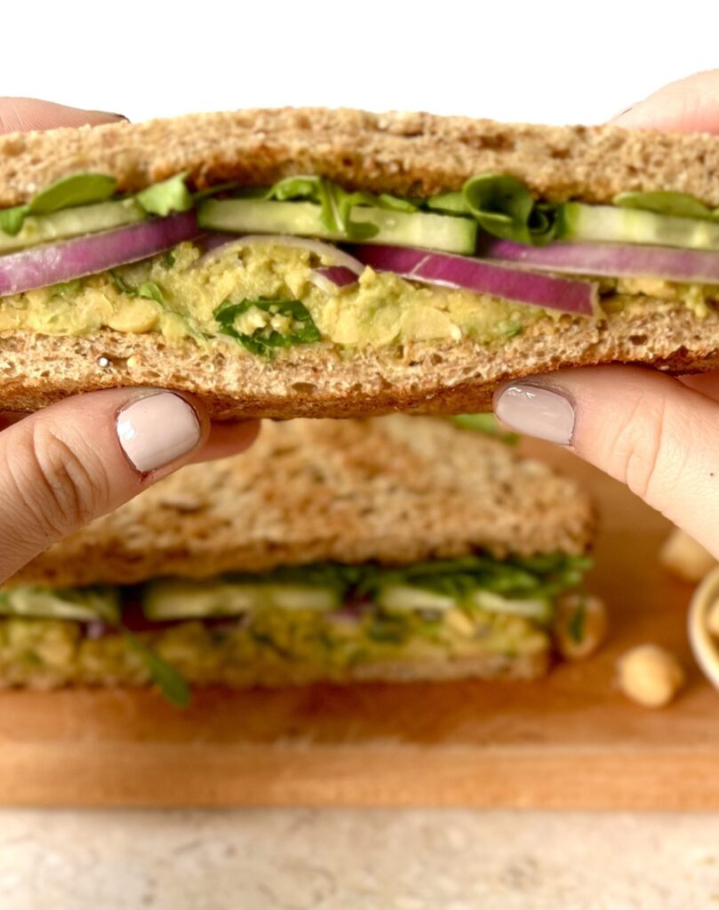 View of the cut side of a avocado chick pea sandwich. There are hands on each side of the sandwich presenting it to the camera.