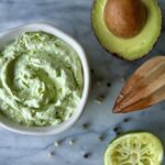 Overhead view of the avocado lime crema in a white bowl. On the side are the avocado and a squished lime.