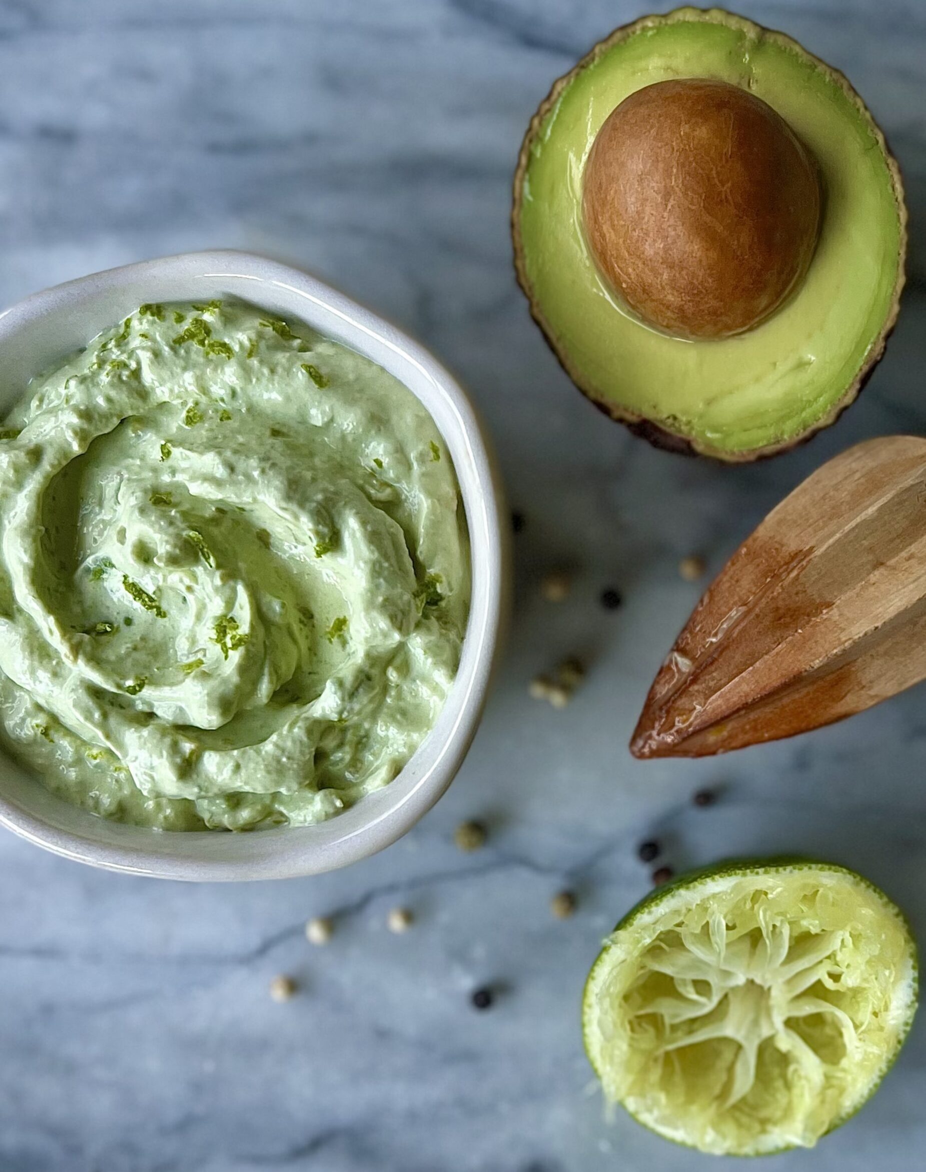 Overhead view of the avocado lime crema in a white bowl. On the side are the avocado and a squished lime.