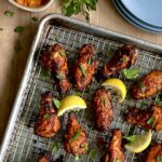 Overhead view of 8 chicken drumsticks and wings with a red coating on a baking sheet with lemon wedges surrounding them.