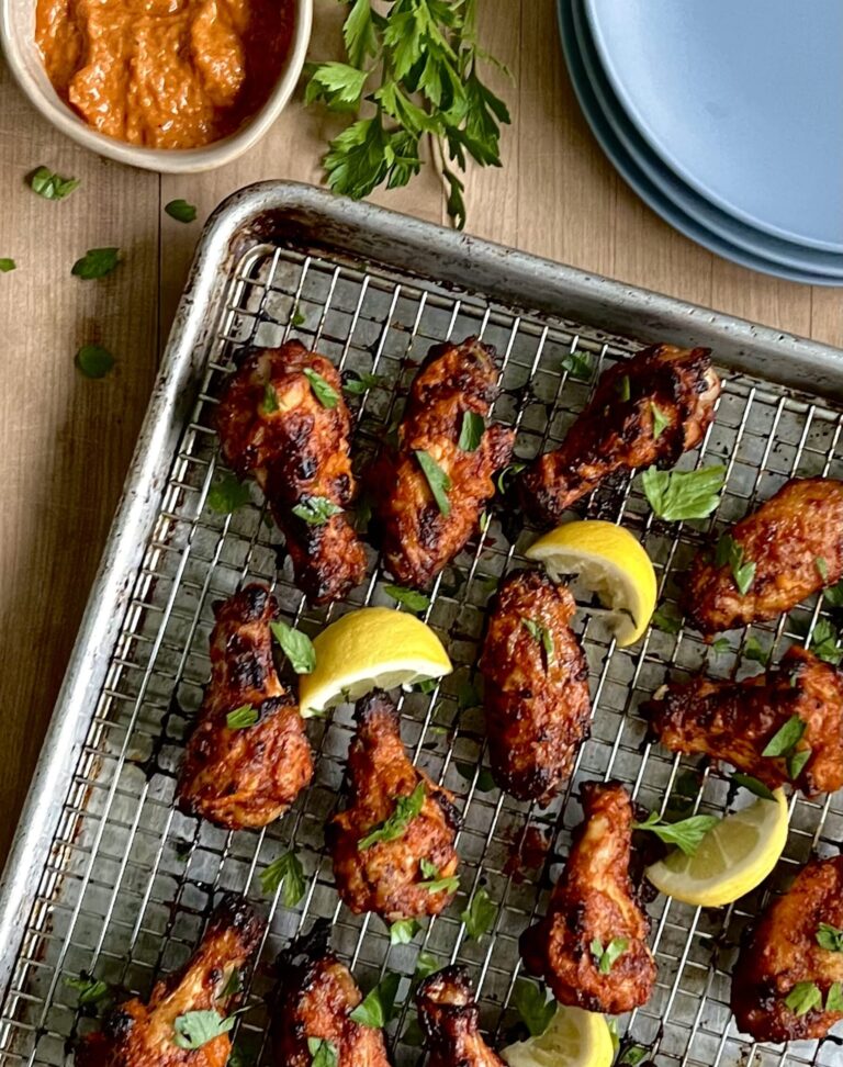 Overhead view of 8 chicken drumsticks and wings with a red coating on a baking sheet with lemon wedges surrounding them.