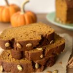 Side view of three thick slices of pumpkin bread stacked on a piece of parchment paper. Two small pumpkins are in the background.