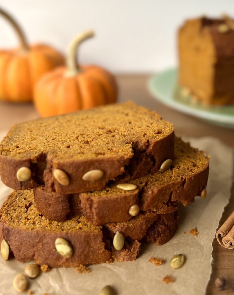 Side view of three thick slices of pumpkin bread stacked on a piece of parchment paper. Two small pumpkins are in the background.