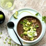 Picture of a bowl of black bean soup from above, topped with cheese, avocado, sour cream, cilantro and green onion. On the side is a bowl of limes.