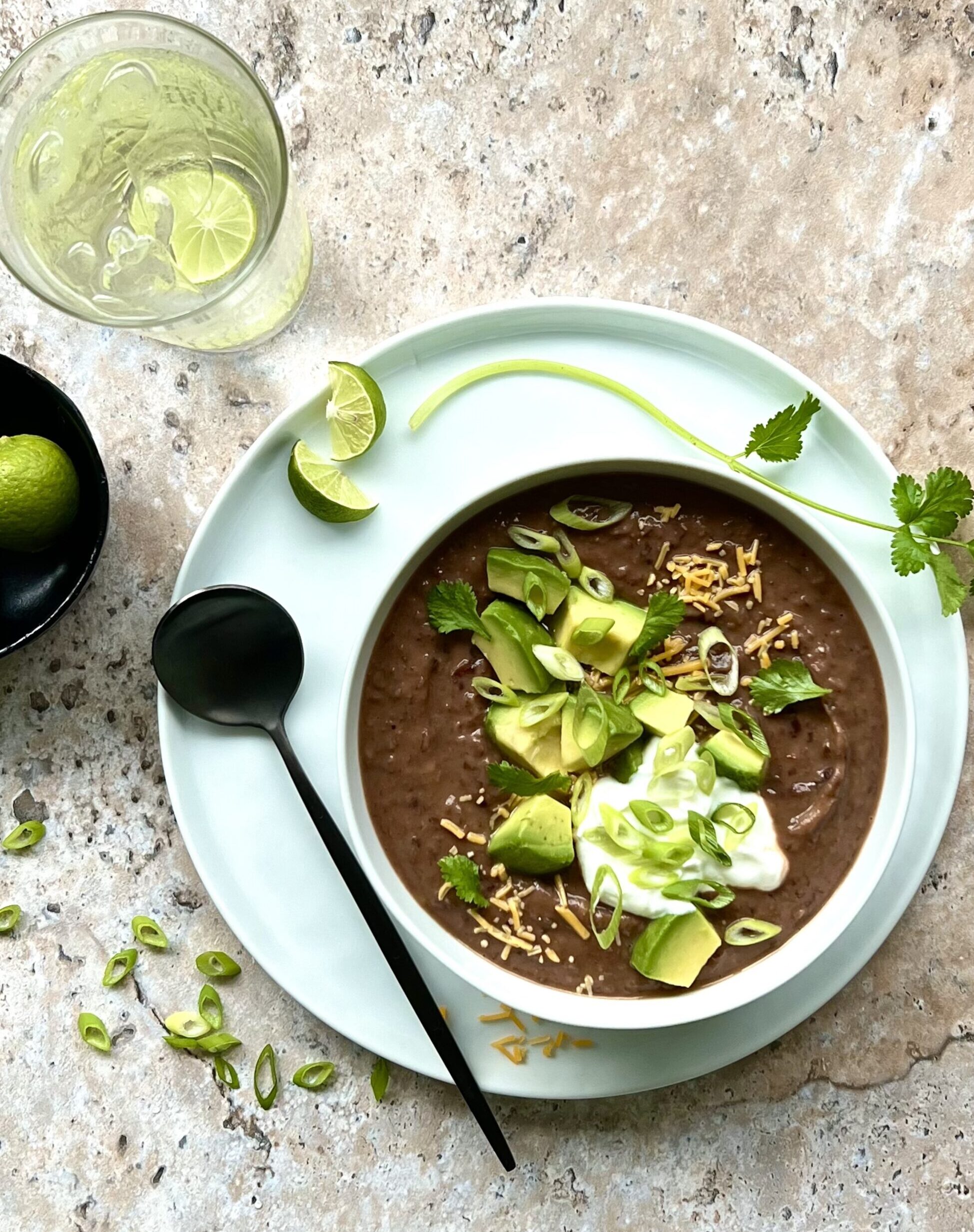 Picture of a bowl of black bean soup from above, topped with cheese, avocado, sour cream, cilantro and green onion. On the side is a bowl of limes.