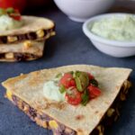 3/4 overhead view of a wedge of the corn and black bean quesadilla. On top of the quesadilla is sour cream, tomatoes, jalepeno. It is on a blue background.