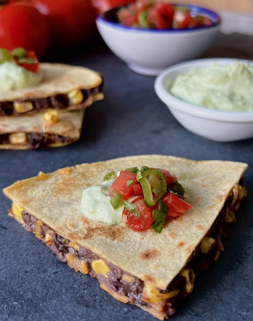 3/4 overhead view of a wedge of the corn and black bean quesadilla. On top of the quesadilla is sour cream, tomatoes, jalepeno. It is on a blue background.