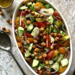 Overhead view of a chopped salad in a white serving dish. Colors of tomatoes, cucumber and chickpeas. There is a spoon on the left side for added effect.