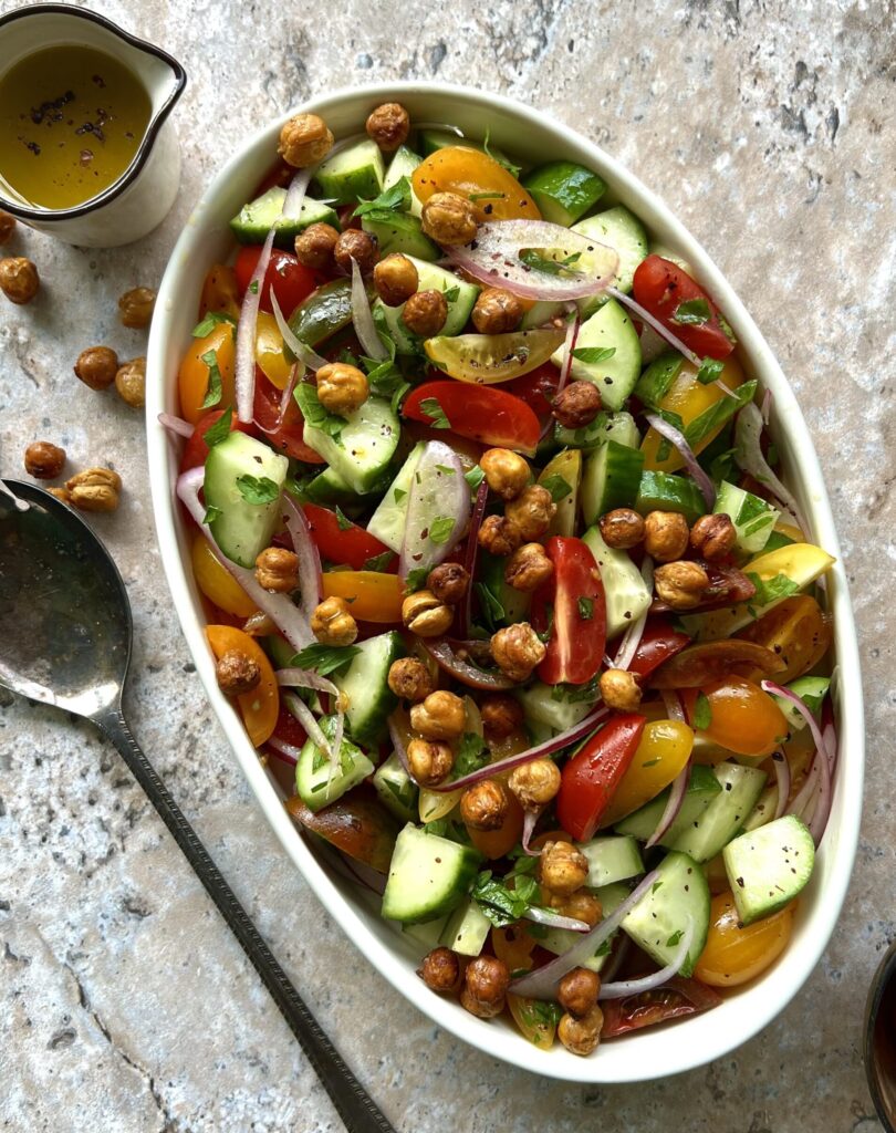 Overhead view of a chopped salad in a white serving dish. Colors of tomatoes, cucumber and chickpeas. There is a spoon on the left side for added effect.