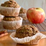 Side view of three muffins. They are in a white wrapper. The one in front is on a brown table top and the ones in the back ground are on a cooling rack and there is an apple on the side for added color.