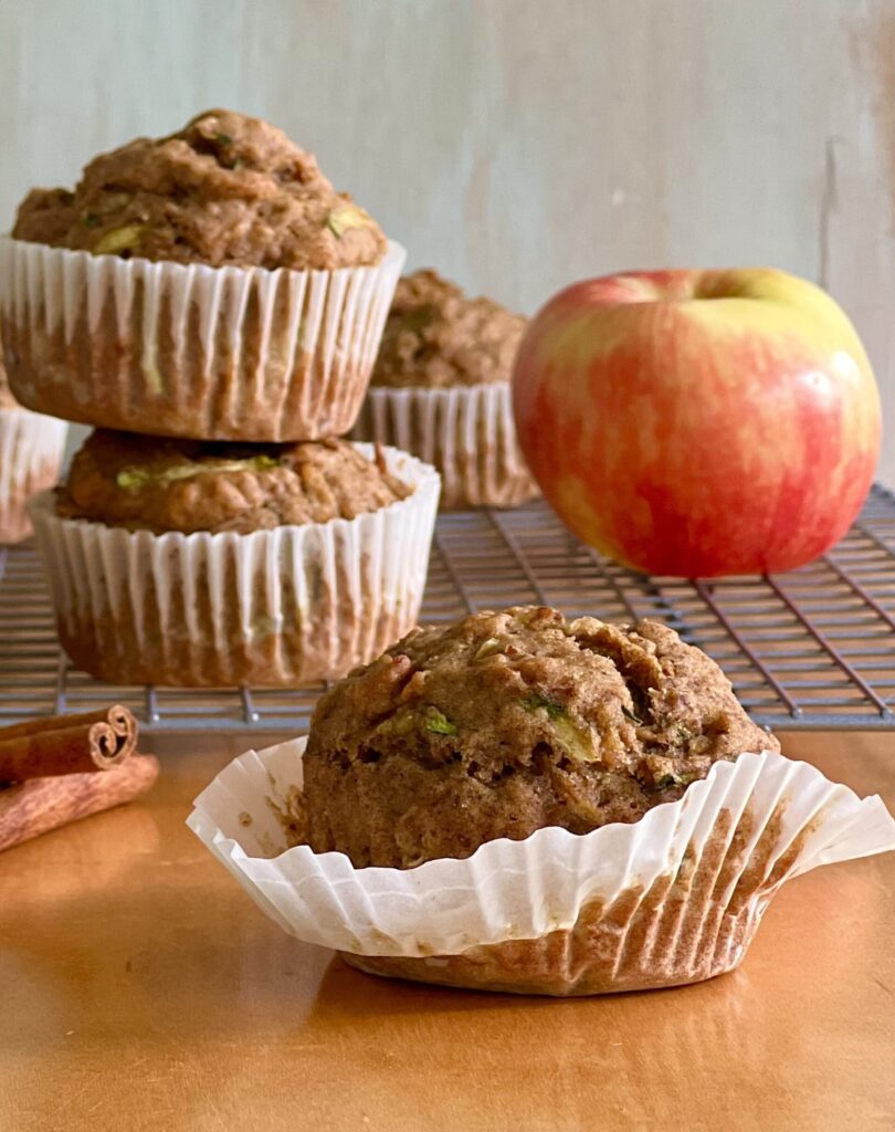 Side view of three muffins. They are in a white wrapper. The one in front is on a brown table top and the ones in the back ground are on a cooling rack and there is an apple on the side for added color.