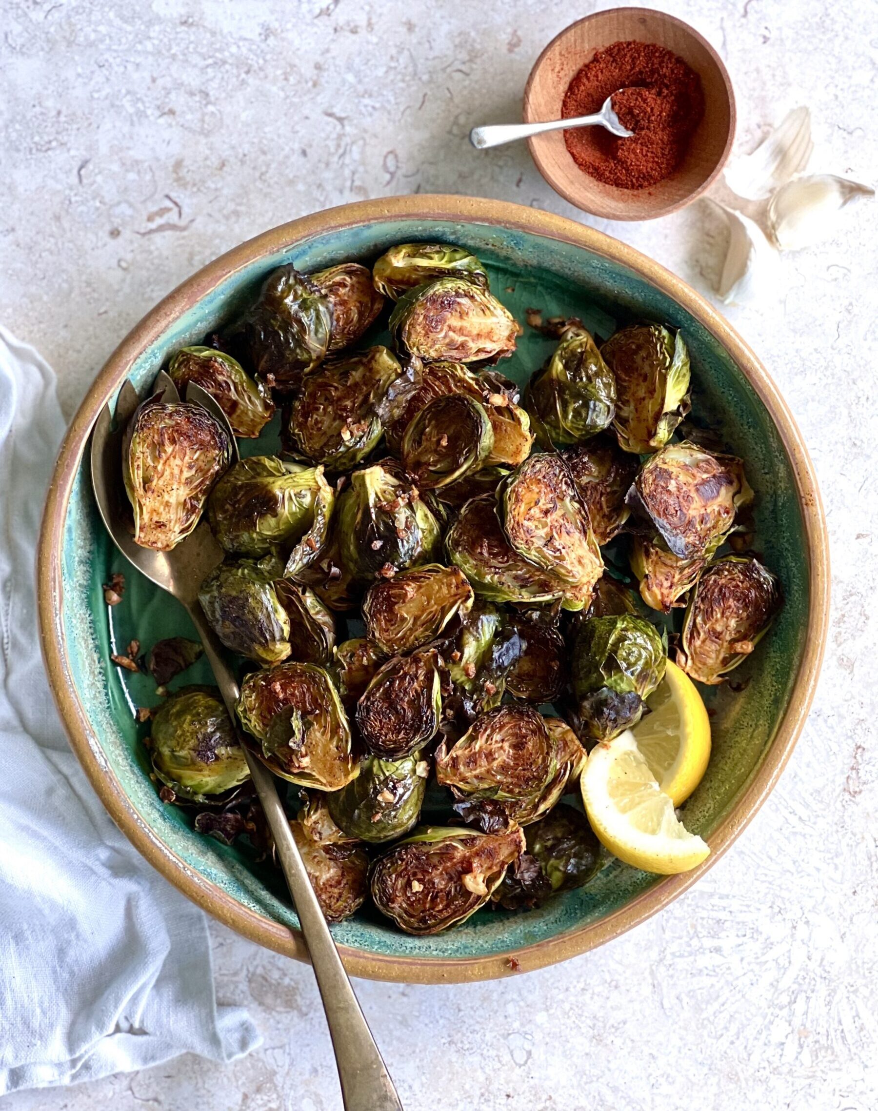 Overhead view of roasted brussel sprouts in a blue bowl with a brown rim. It is on a white marble background with a small bowl of paprika on the side.