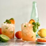 Side view of two short glasses filled with ice and an orange drink. There is basil draped along the top of the glasses and a half slice of orange. In the background are a lemon, a lime and an empty glass bottle.