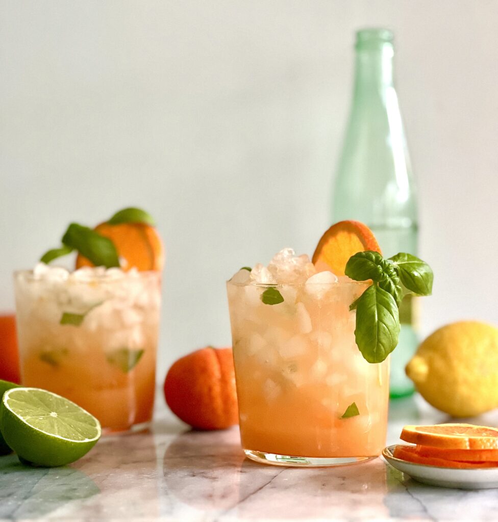 Side view of two short glasses filled with ice and an orange drink. There is basil draped along the top of the glasses and a half slice of orange. In the background are a lemon, a lime and an empty glass bottle.