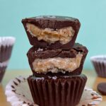 Side view of 3 stacked chocolate almond butter cups. The top to are broken in half so you can see the inside of the almond butter. It is set against a blue background.