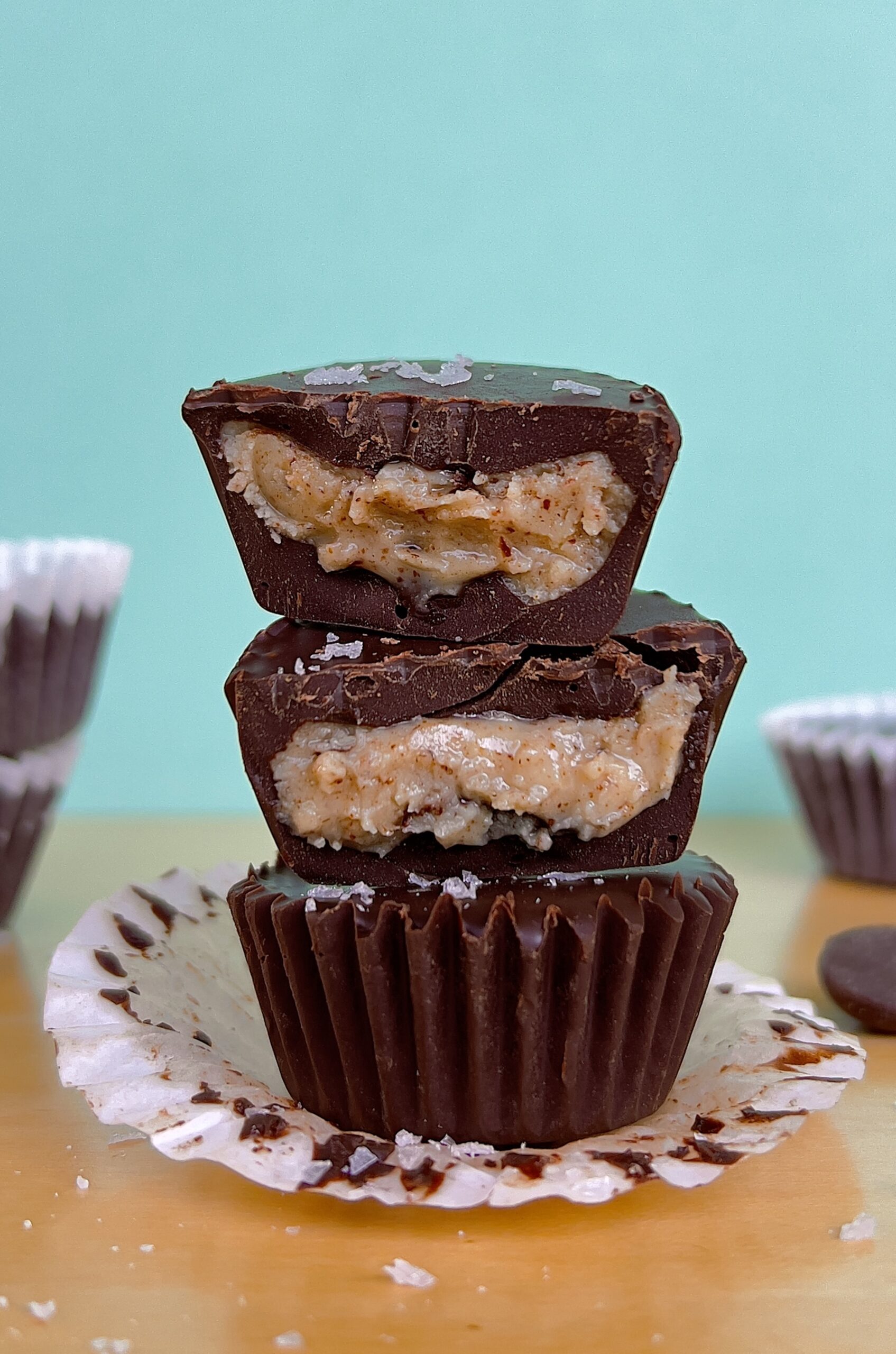 Side view of 3 stacked chocolate almond butter cups. The top to are broken in half so you can see the inside of the almond butter. It is set against a blue background.