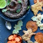 Blue bowl in the upper left corner with black bean dip swirled inside of it. It is topped with parsley and alongside of the bowl are chips made in the shape of ghosts, bats and pumpkins.