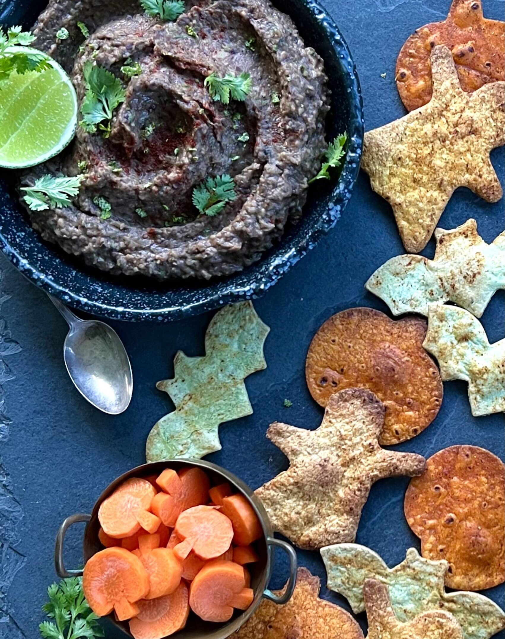 Blue bowl in the upper left corner with black bean dip swirled inside of it. It is topped with parsley and alongside of the bowl are chips made in the shape of ghosts, bats and pumpkins.