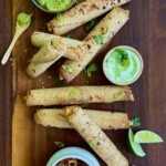 Overhead view of chicken taquitos on a dark wooden cutting board with a small bowl of avocado crema on the right and two lime wedges.