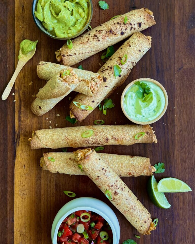 Overhead view of chicken taquitos on a dark wooden cutting board with a small bowl of avocado crema on the right and two lime wedges.