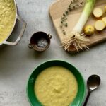 Overhead view of yellow squash soup in a green bowl with a cutting board in the upper right that has squash, leeks and thyme on it.