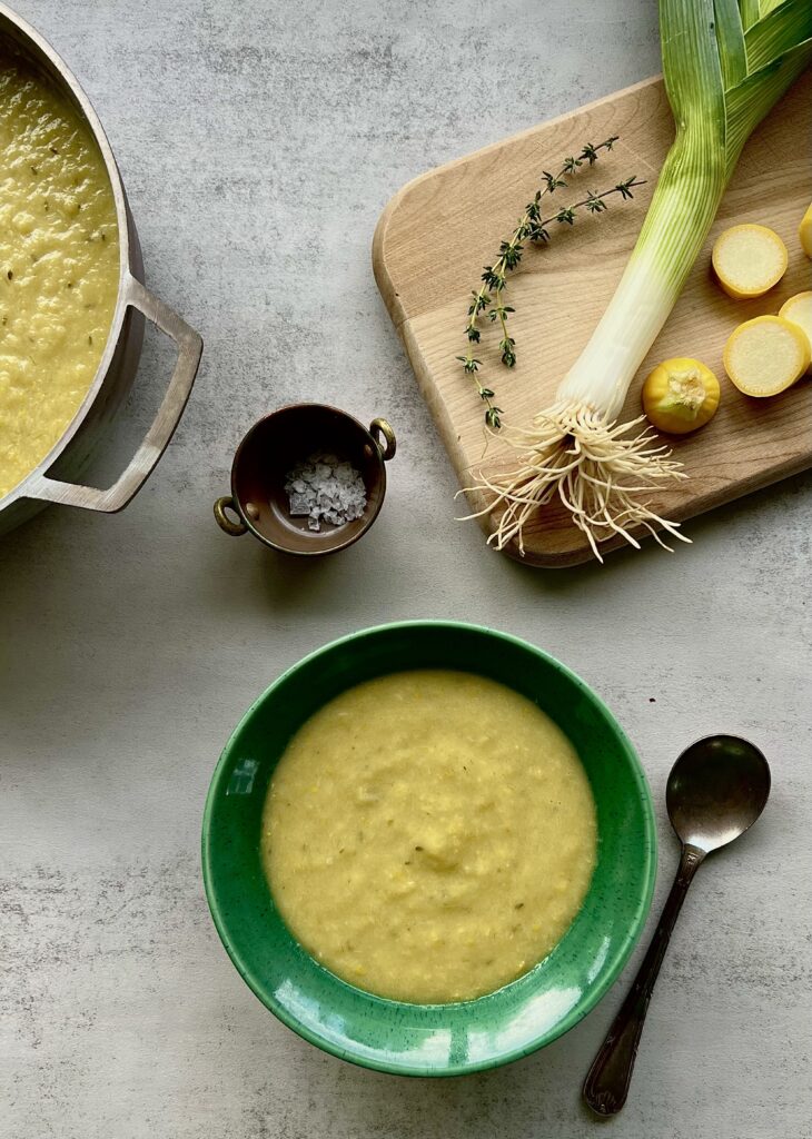 Overhead view of yellow squash soup in a green bowl with a cutting board in the upper right that has squash, leeks and thyme on it.