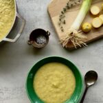 Overhead view of yellow squash soup in a green bowl with a cutting board in the upper right that has squash, leeks and thyme on it.