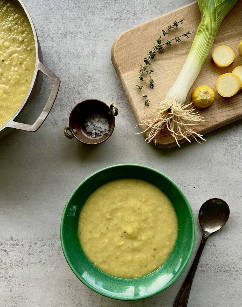 Overhead view of yellow squash soup in a green bowl with a cutting board in the upper right that has squash, leeks and thyme on it.