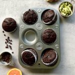 Overhead view of muffin tin with 5 chocolate muffins. The 6th muffin is along the side and there is ginger, shredded zucchini, chocolate chips and an orange slice decorating the background.