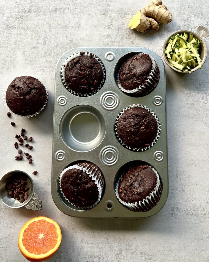 Overhead view of muffin tin with 5 chocolate muffins. The 6th muffin is along the side and there is ginger, shredded zucchini, chocolate chips and an orange slice decorating the background.