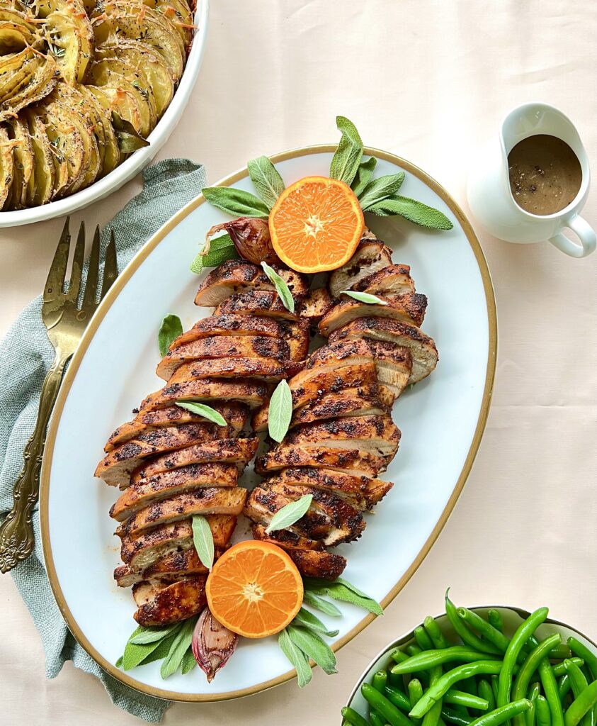 Overhead view of sliced turkey tenderloin on a platter. There is a cut orange on each end and a pitcher of gravy on the side.