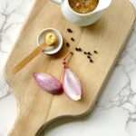 Overhead view of a cutting board with the gravy in a white gravy pitcher sitting on a light brown cutting board. Also on the cutting board are shallots and a teaspoon with dijon mustard.