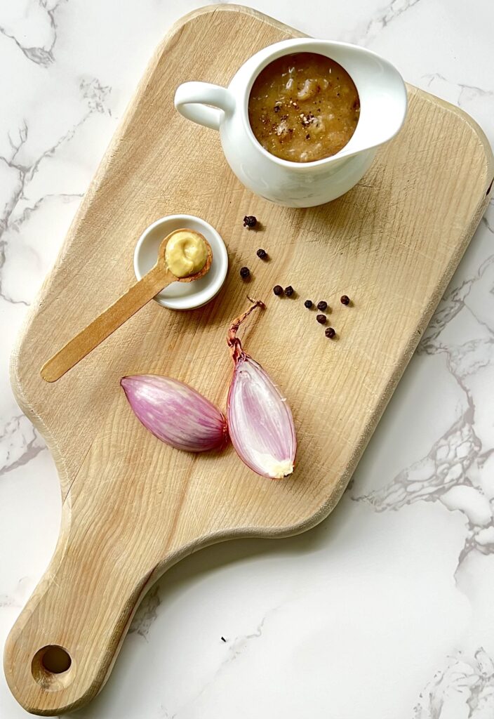 Overhead view of a cutting board with the gravy in a white gravy pitcher sitting on a light brown cutting board. Also on the cutting board are shallots and a teaspoon with dijon mustard.