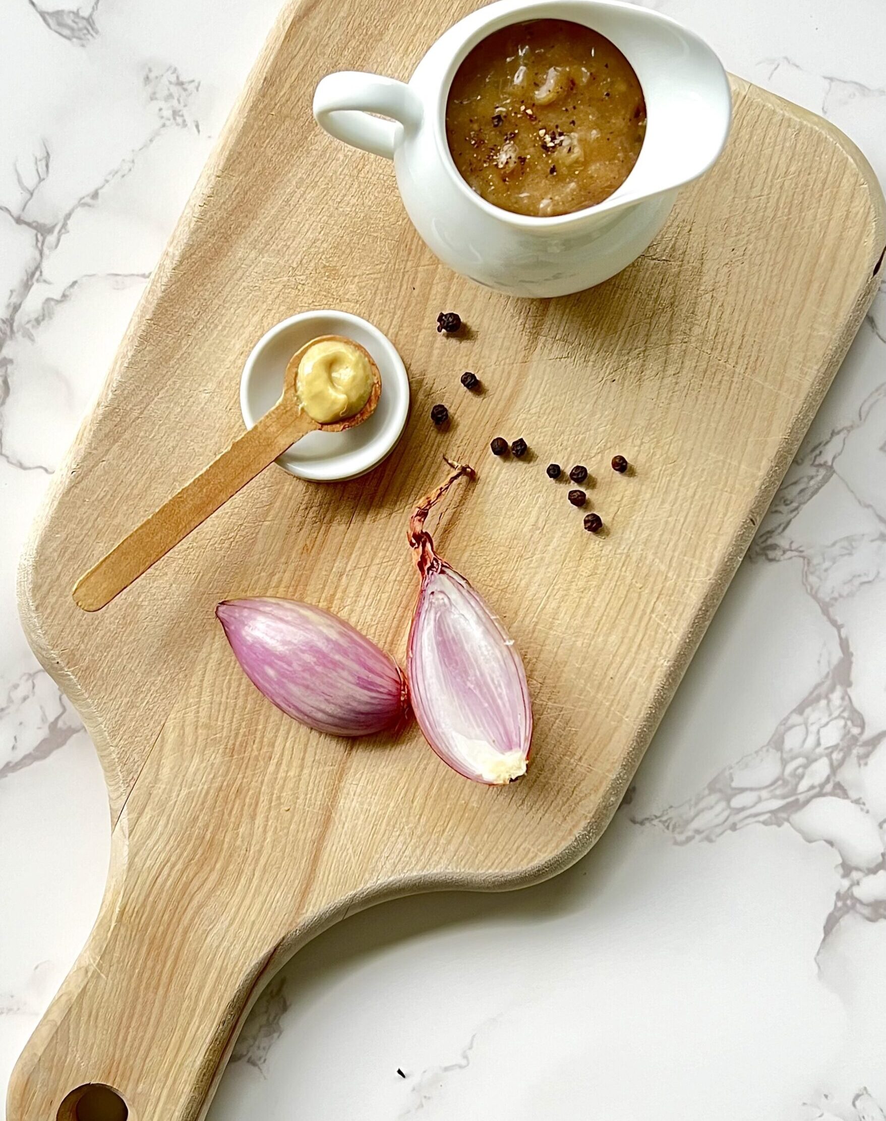 Overhead view of a cutting board with the gravy in a white gravy pitcher sitting on a light brown cutting board. Also on the cutting board are shallots and a teaspoon with dijon mustard.