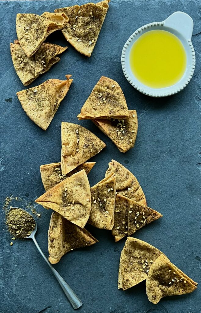 Overhead view of pita chips sprinkled with za'atar seasoning on a blue background with olive oil on the side.