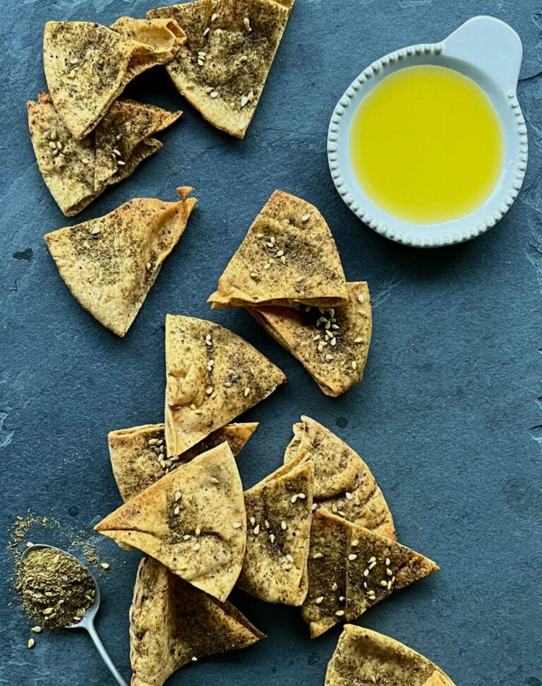 Overhead view of pita chips sprinkled with za'atar seasoning on a blue background with olive oil on the side.