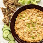 Overhead view of buffalo chicken dip in a burgundy bowl. Celery, pita chips and cucumber are along the side on a white marble background.