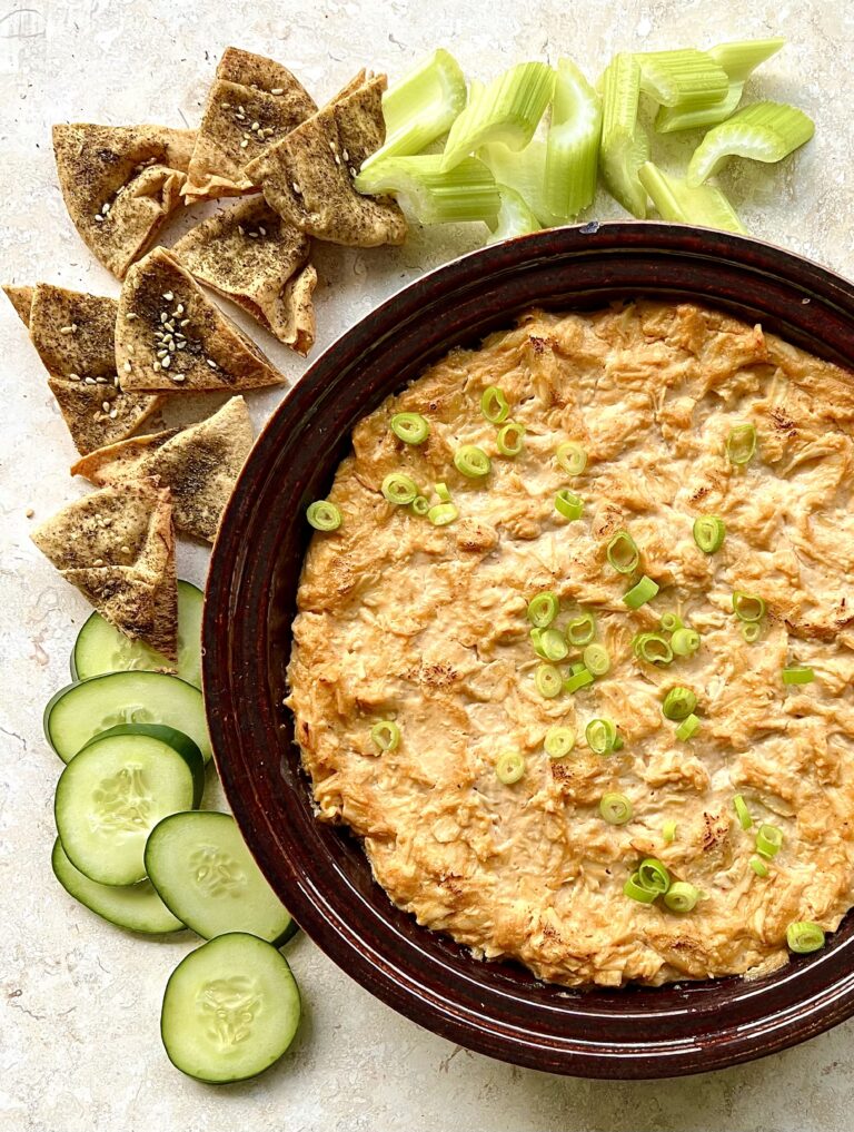 Overhead view of buffalo chicken dip in a burgundy bowl. Celery, pita chips and cucumber are along the side on a white marble background.