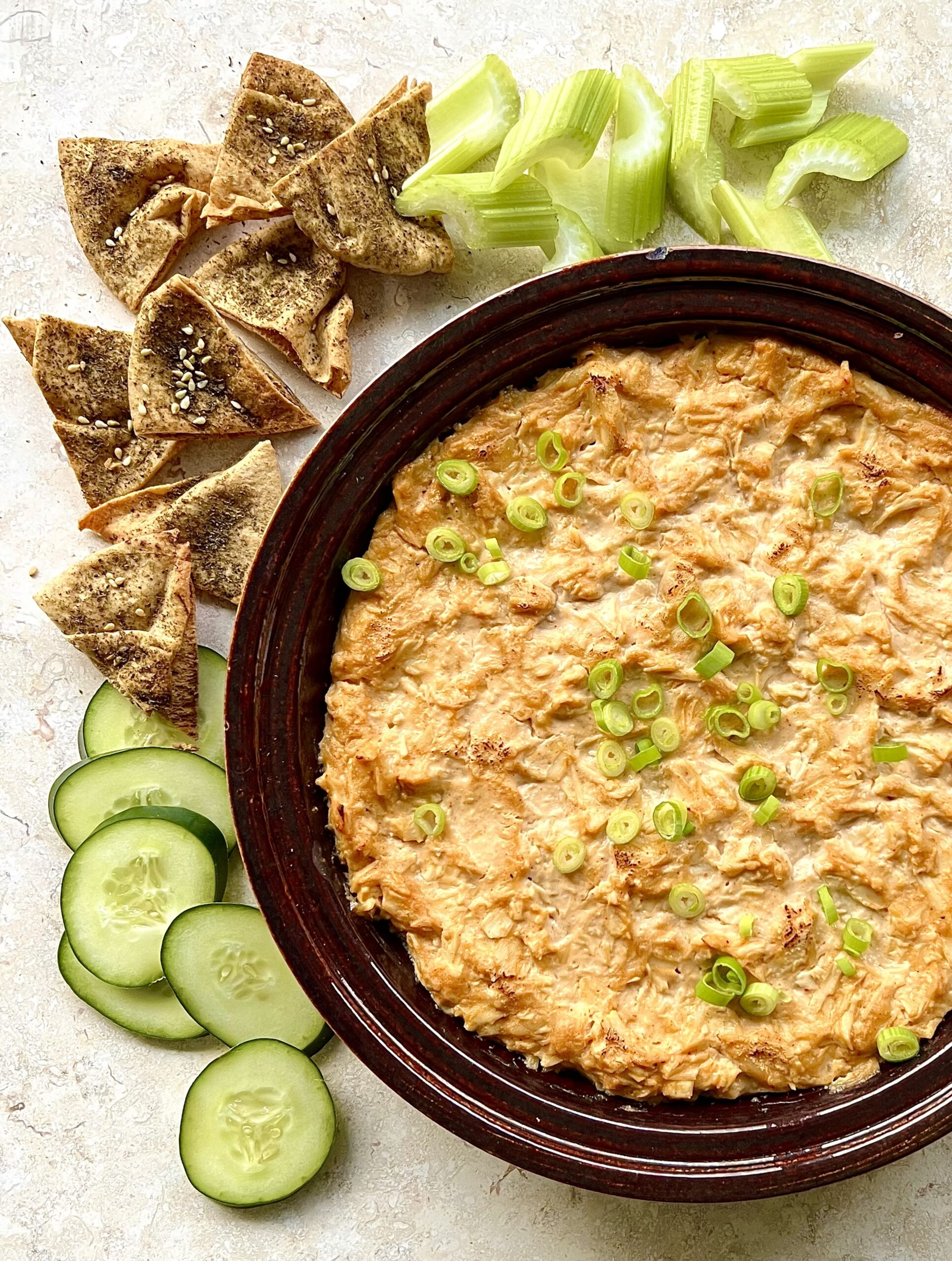 Overhead view of buffalo chicken dip in a burgundy bowl. Celery, pita chips and cucumber are along the side on a white marble background.