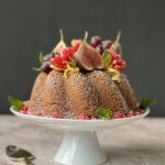 Side view of a bundt cake on a white cake stand. The cake is topped with fruit and powdered sugar.