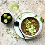 Picture of a bowl of black bean soup from above, topped with cheese, avocado, sour cream, cilantro and green onion. On the side is a bowl of limes.