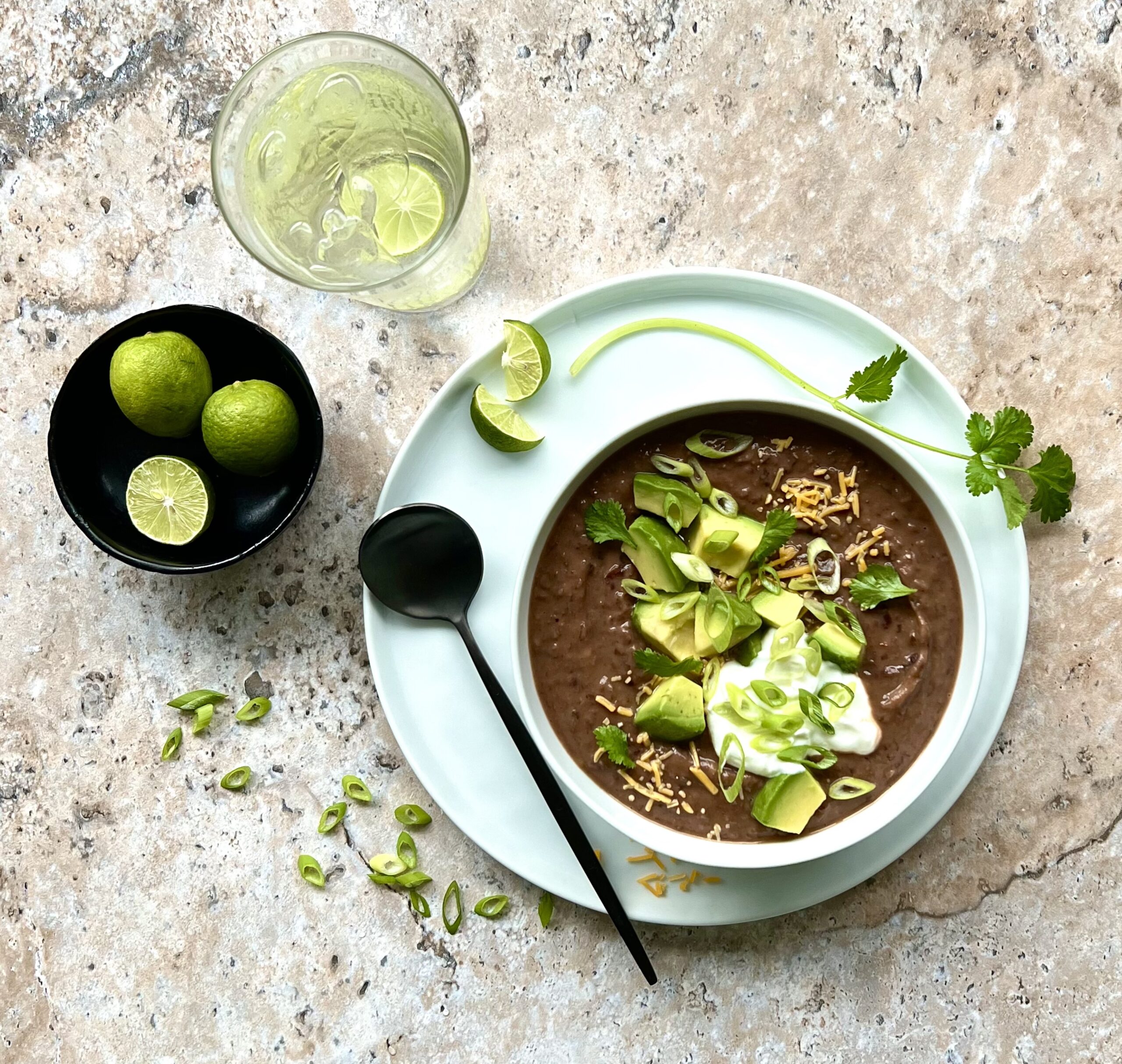 Picture of a bowl of black bean soup from above, topped with cheese, avocado, sour cream, cilantro and green onion. On the side is a bowl of limes.