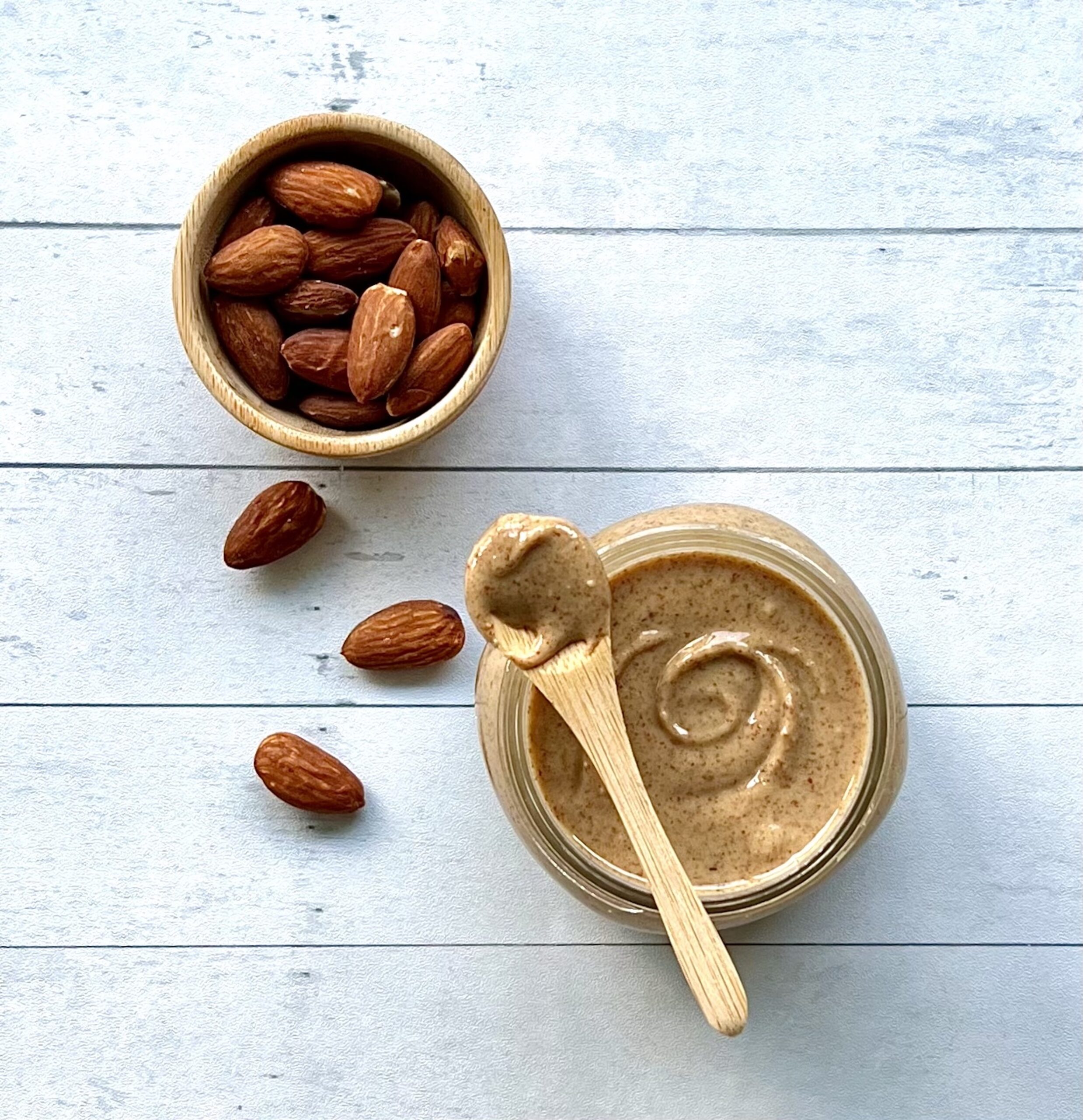 Overhead shot of a cup of almonds and next to it is a cup of swirled almond butter with a wooden spoon with a spoonful of almond butter resting on top of the jar.