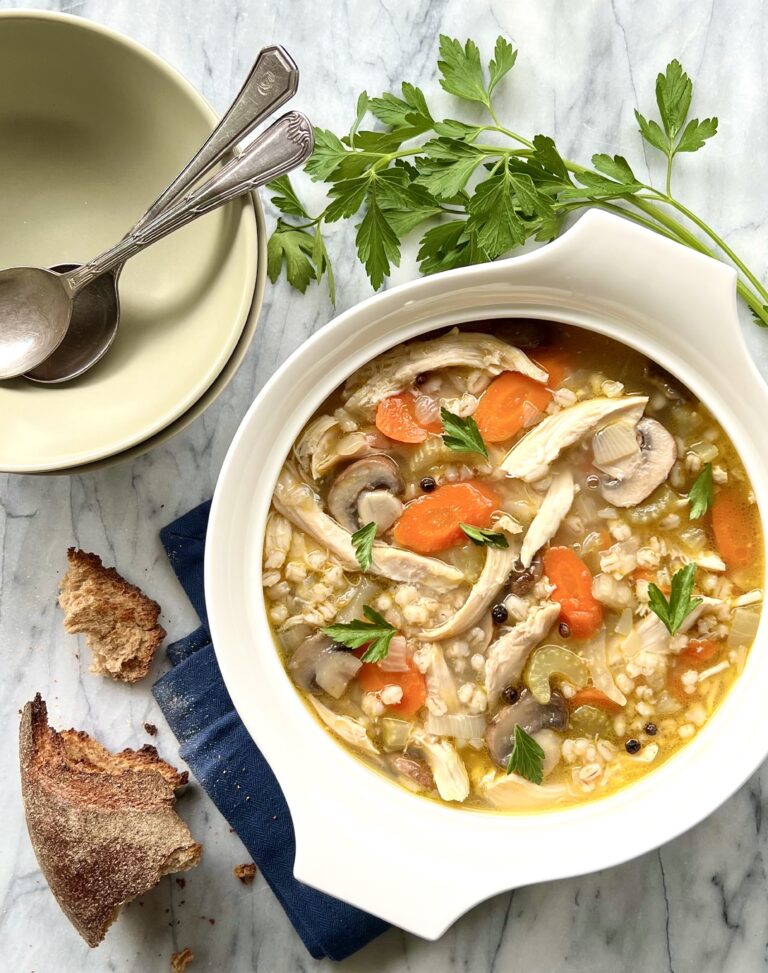 Overhead view of chicken mushroom barley soup. In a white bowl on a marble background with parsley and toasted bread adorning the background.