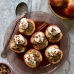 Overhead shot of 5 braised half apples topped with granola and greek yogurt. The apples are resting on a wooden dish with a rustic spoon draped across the left side of the dish.