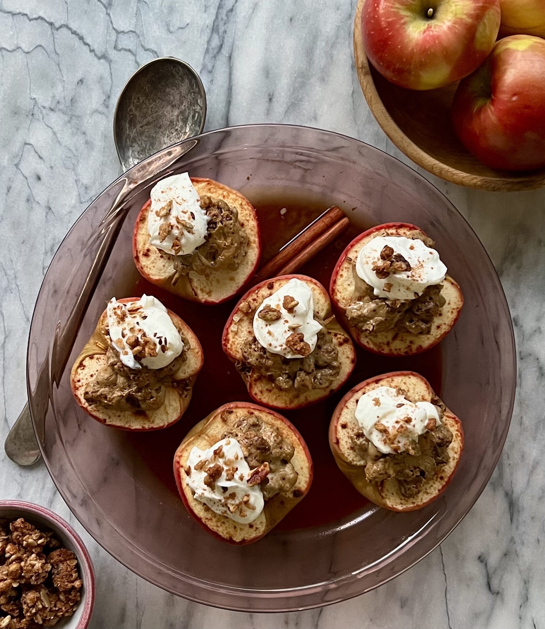 Overhead shot of 5 braised half apples topped with granola and greek yogurt. The apples are resting on a wooden dish with a rustic spoon draped across the left side of the dish.