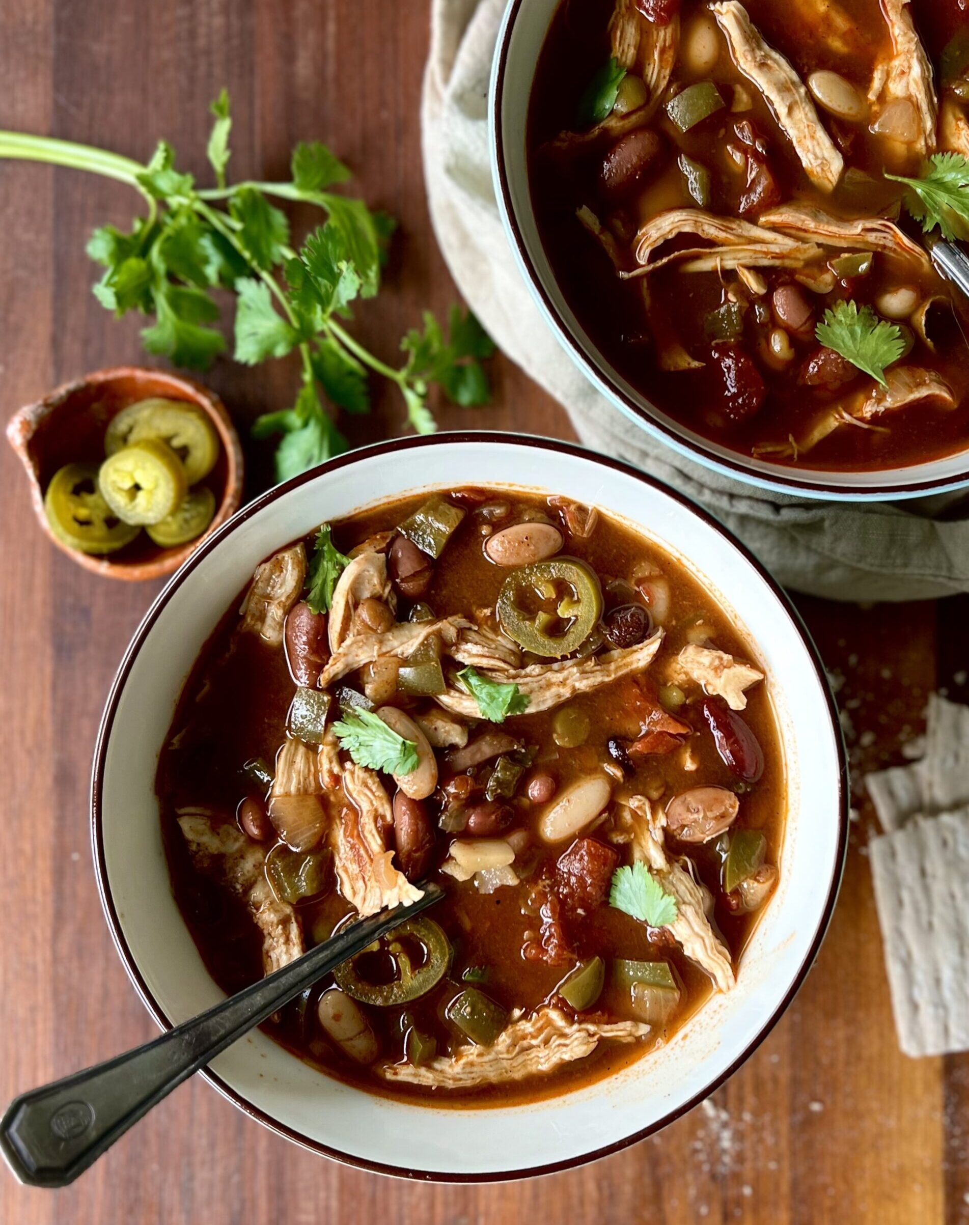 Overhead view of two bowls of bean soup on a wooden cutting board background and a piece of parsley and a cup of jalepenos in the background.