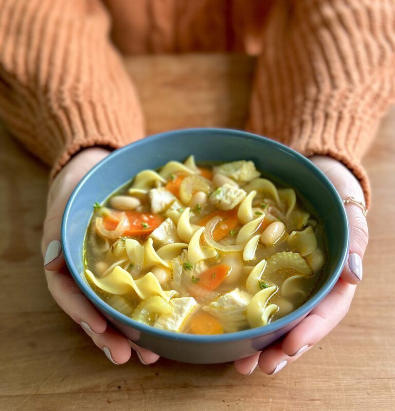 Chicken Noodle Soup in a blue bowl being held by two hands with light nail polish and an orange sweater. It is presented to the camera.