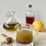 The dressing is pictured in a glass cup with a handle and a lip to pour. In the background are a container of olive oil, vinegar and a lemon. To the side is a wooden teaspoon of sumac.